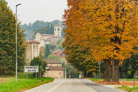 Autumn scene in Monteu da Po, a charming village in the Piedmont region of Italy with a quiet country road lined with colorful fall trees, leading toward historic buildings and acの写真素材