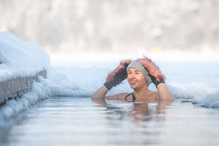 Vilnius, Lithuania - January 11 2026: Smiling pretty girl wearing mittens and hat immersed in the freezing cold water of a lake practicing cold water swimming. Wim Hof Method, coldのeditorial素材