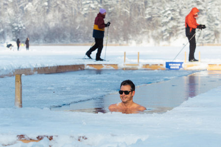 Vilnius, Lithuania - January 11 2026: Man or boy wearing sunglasses immersed in the freezing cold water of a lake practicing cold water swimming with skiers in the background. Wimのeditorial素材