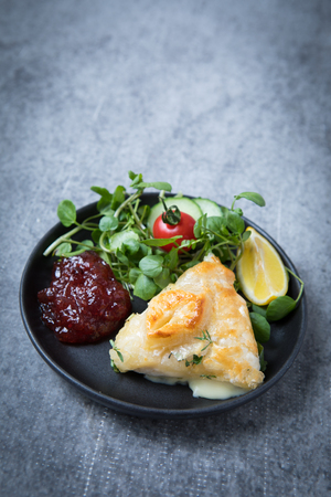 Camembert en croute cheese in puff pastry served on a black plate with red jam, salad, vegetables and lemon wedge on a grey grunge background.の写真素材