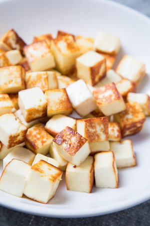 Fried small cubes of paneer in the foreground on a white plate. Close up picture of small fried cubes of cottage cheese. Pan-fried paneer cubes.の写真素材