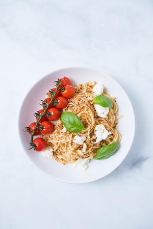 Spaghetti pasta with breadcrumbs, mozarella, fresh basil and a vine of tomatoes in a deep white plate with a light background. Italian pasta dish with mozarella and tomatoes.の写真素材