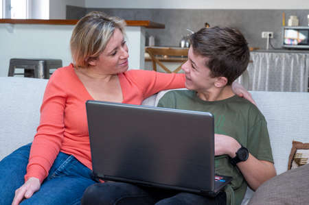 Blonde mom sitting on sofa with teenage son helping with homework online at computerの写真素材