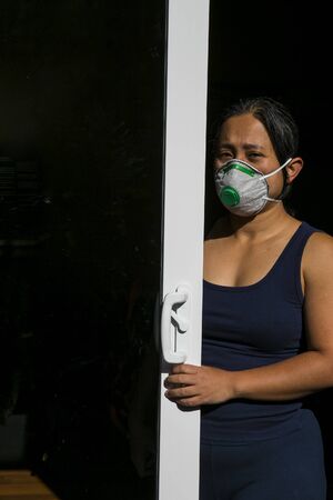 A concerned woman stands behind glass door with face mask. Suitable to depict self-quarantine at...