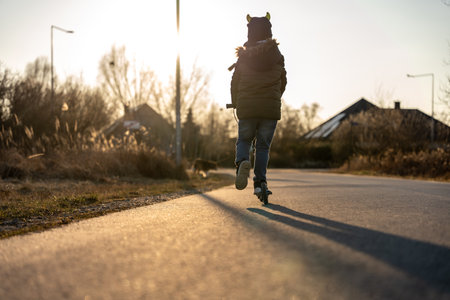Child riding scooter alone down road at sunset in quiet neighborhood showing childhood adventure and...