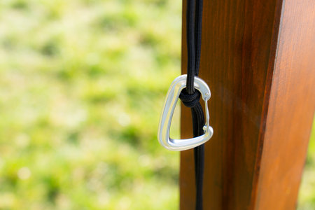 Silver carabiner clip tied with black rope on wooden frame, with green blurred grassy background,...