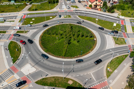 Large roundabout intersection with greenery and road markings surrounded by vehicles and urban roadsの写真素材
