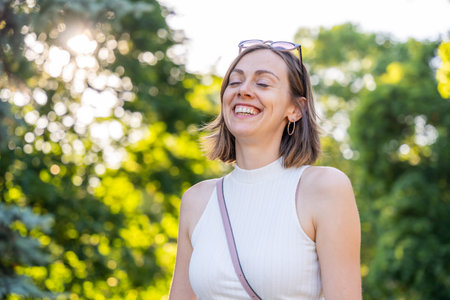 Laughing woman in white sleeveless top with closed eyes enjoying summer light in green park, casual relaxed outdoor lifestyleの写真素材