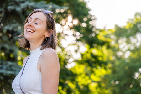 Smiling woman laughing in sunny green park with trees in background, joyful and bright atmosphere during warm summer dayの写真素材
