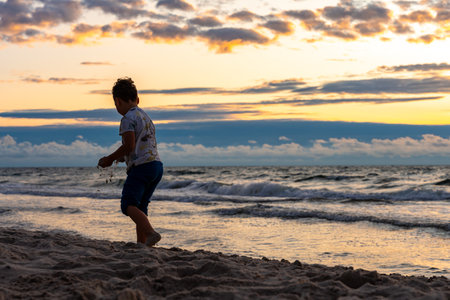 Child playing on sandy beach at sunset near waves, silhouetted figure in warm evening light, carefree summer momentの写真素材