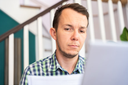 Man Reading Paper Documents While Using Laptop At Home Office Remote Workの写真素材