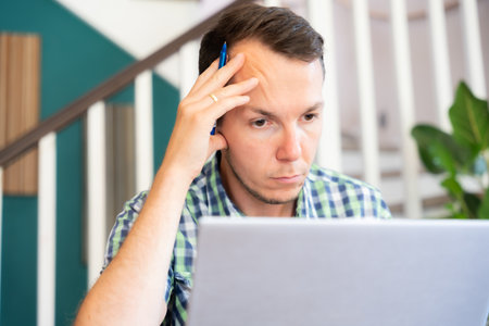Focused Man Holding Pen Looking At Laptop Screen While Working Remotely At Home Officeの写真素材