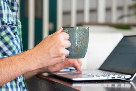 Person Holding Coffee Mug While Working On Laptop Indoors For Remote Work And Home Office Productivityの写真素材