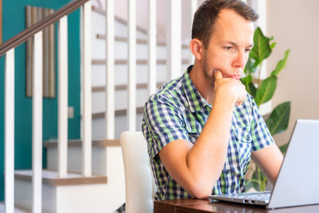 Serious Man Holding Chin Thinking While Looking At Laptop Screen At Home Officeの写真素材