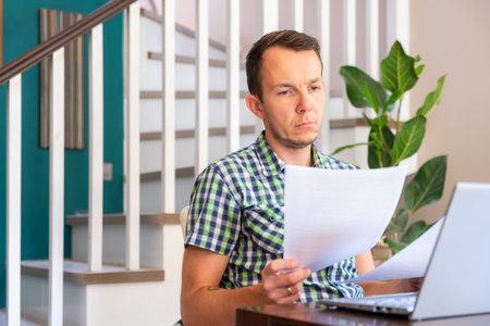 Man Holding Paper Documents While Working With Laptop At Home Officeの写真素材