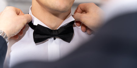 Best Man Adjusting Groom Black Bow Tie On White Shirt During Elegant Wedding Preparationの写真素材
