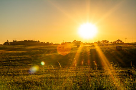 Golden Sunrise Over Rural Field With Sun Rays And Hay Bales In Countryside Meadow, Bright Agricultural Grass Landscape Backgroundの写真素材