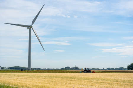 A farmer with a tractor works his arable land on the land is a large windmill and the sky is blue