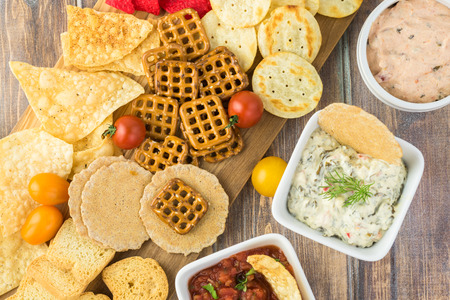 Top view of cutting board with variety of snacks and bowls with dip and salsa.