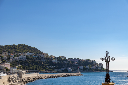 View over a bay in front of Nice, France, with a crucifix in the foreground. In the background you can see buildings and a protective wall with a lighthouse.の写真素材