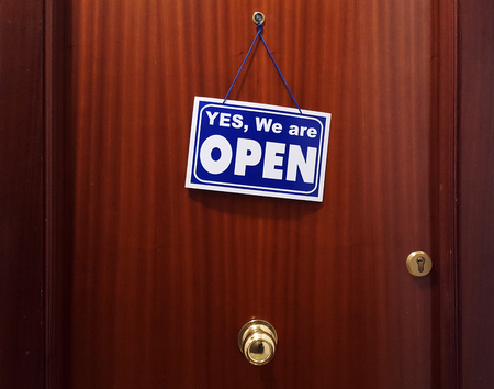 Brown flat door with a sign saying Yes We Are Open. Spanish building.