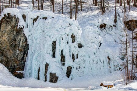 Stunning winter landscape with a house surrounded by forest at the foot of cliffs and frozen waterfallの写真素材