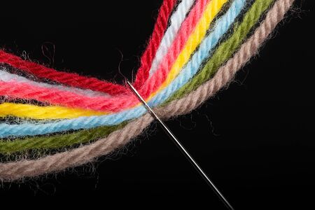 On a black background, several bright multicolored woolen threads are pass through the eye of the...