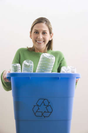 An attractive young woman his smiling and holding a blue recycle bin with plastic bottles in it. Vertical shot.の写真素材