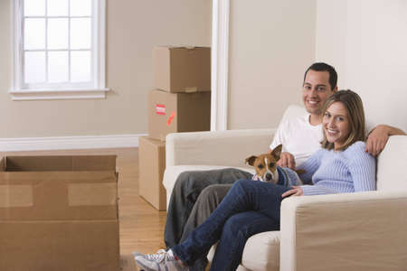 A young couple and their dog are sitting on a couch in a living room. Moving boxes are arranged on the floor around them. Horizontal shot.の写真素材