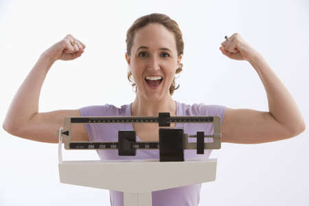 A young woman flexes her arms while standing on a scale and smiling at the camera. Horizontal shot. Isolated on white.の写真素材