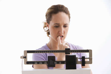 A young woman is standing on her scale and checking her weight. She has a worried expression on her face. Horizontal shot. Isolated on white.の写真素材