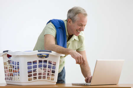 A man is working on a laptop while he folds laundry.  He is smiling and looking away from the camera.  Horizontal shot.の写真素材