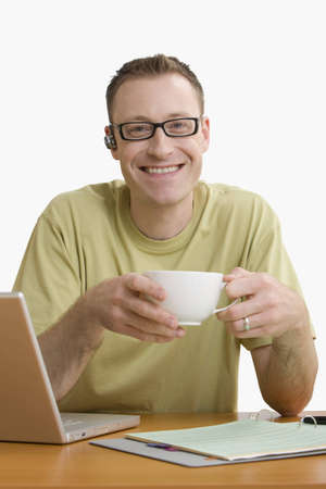 Man with a cellphone ear piece and sitting at his desk enjoys a cup of coffee while smiling for the camera.  Vertical shot.  Isolated on white.の写真素材