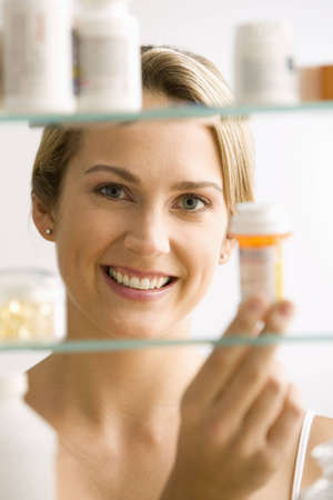 A young woman looks through a medicine cabinet and smiles at the camera.  Vertical shot.の写真素材