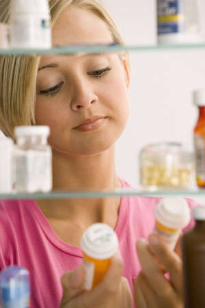 A young woman is reading the labels of to medicine containers from her medicine cabinet.  Vertical shot.の写真素材