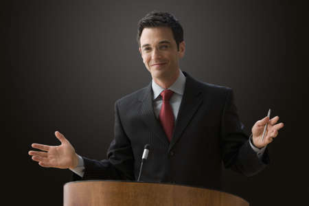 A businessman is standing at a podium with a microphone giving a lecture with outstretched hands. Horizontal shot.の写真素材