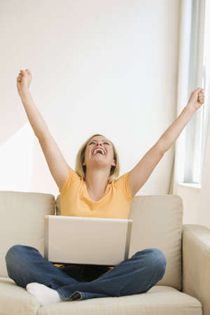 A young woman sitting on the couch with a laptop throws her arms in the air while cheering.  Vertical shot.の写真素材