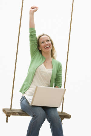 A young woman is sitting on a swing while working on a laptop.  She is raising a fist into the air.  Vertical shot.の写真素材