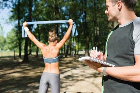Personal fitness trainer with client, resistance band exercise, focus on hand and notebook