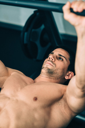 Muscular man lifting weights, lying on weight bench gym. Focus on face, color graded imageの写真素材