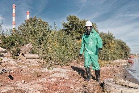 Environmentalist in protective suit working at a pollution site. Contamination of air, water and soil. Pollution factors like factory chimneys, old tires, plastic bottles, chemicals and other waste, all represented in the photo.の写真素材