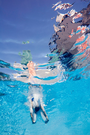 Professional lady diver breaks the surface after backfilp dive from a springboard. Interesting reflections of sky and diving tower in the upper part. Underwater wide angle.の写真素材