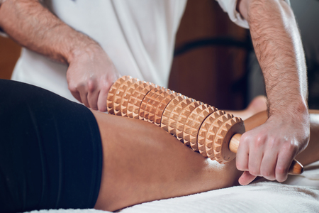 Close up of anti-cellulite cellulite Madero therapy hand massage with wooden rolling pin, beautiful young sporty woman lying on white towel during treatmentの写真素材
