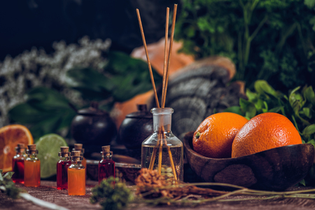 Bottles filled with red and orange essential oils and aromatherapy bottle with wooden sticks inside. Fresh citrus fruit, orange and lime cut in half.  Green plants background.の写真素材