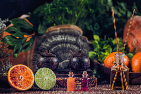 Bottles filled with red and orange essential oils and aromatherapy bottle with wooden sticks inside. Fresh citrus fruit, orange and lime cut in half.  Green plants background.の写真素材