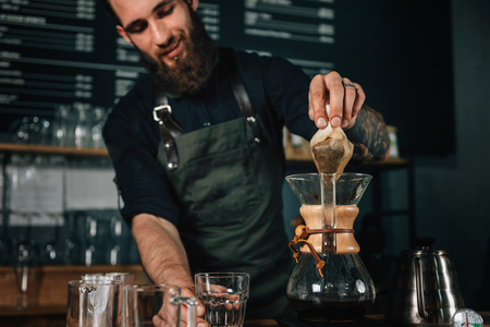 Portrait of young male barista smiling. Barista with tattooed arms wearing dark uniform. Tools and...