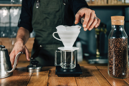 Male barista hands putting white filter to ceramic dripper. Tools and equipment for making Drip Brew coffee on wooden table.  Barista with tattooed arms wearing dark uniform.の写真素材