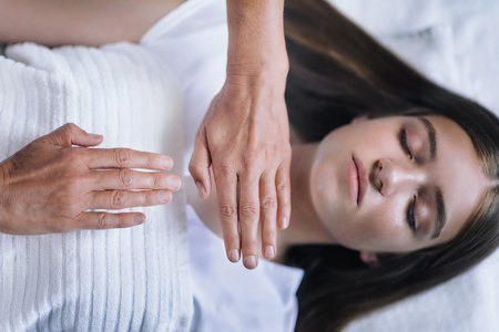Beautiful teenage girl in Reiki spiritual healing session. Reiki therapist holding hands above heart and throat chakra and transfer energy. Peaceful teenage girl lying with her eyes closed.の写真素材