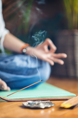 Woman meditating in lotus position on turquoise yoga mat on wooden floor. Focus on incense stick and smoke. Unrecognizable yoga practitioner in the background. Relax after Yoga trainingの写真素材