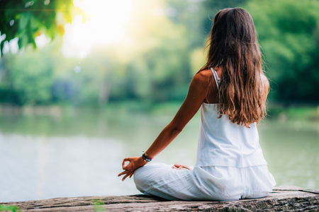 Woman doing yoga by the lake, sitting in lotus pose with fingers in mudra position.の写真素材